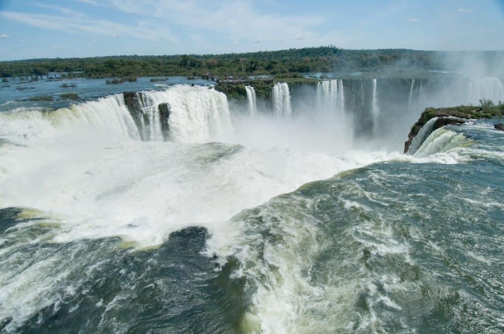 Imagem da Garganta do Diabo, a maior e mais impressionante das quedas das Cataratas do Iguaçu. A poderosa queda d'água desce em um formato de ‘U’, formando uma densa névoa que se eleva no ar devido à força da água caindo de aproximadamente 80 metros de altura. A correnteza corre vigorosamente, enquanto a vegetação densa cobre as margens ao fundo, criando um contraste entre o verde das plantas e o branco turbulento das águas. O céu claro e azul destaca ainda mais a grandiosidade e beleza natural deste ponto icônico.