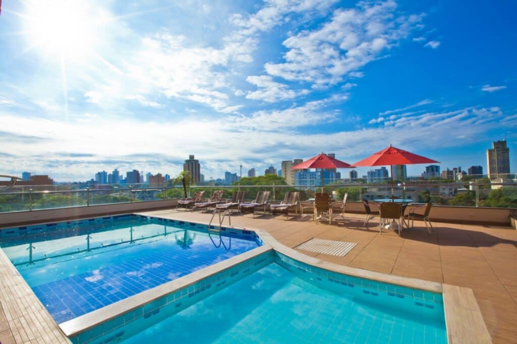 Área externa de um hotel em Foz do Iguaçu, destacando uma piscina aquecida com espreguiçadeiras ao redor, guarda-sóis vermelhos e uma vista panorâmica da cidade, ideal para quem busca hotéis com piscina aquecida em Foz do Iguaçu.