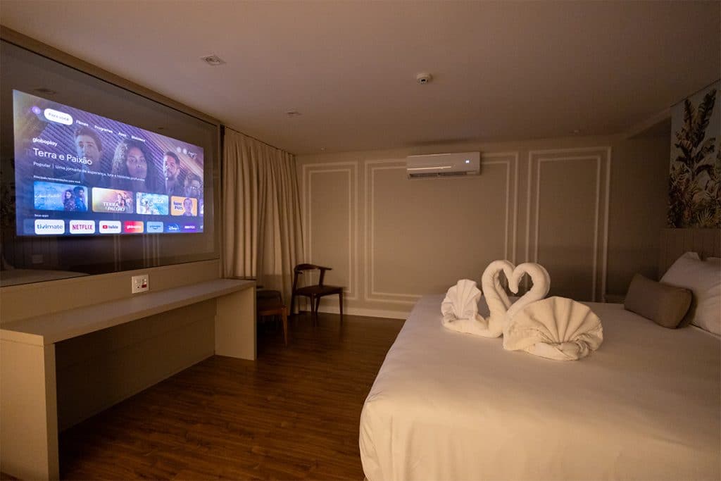 Suíte aconchegante do Del Rey Quality Hotel, com cama de casal decorada com toalhas em forma de cisnes. O ambiente moderno possui uma TV de tela grande exibindo plataformas de streaming e iluminação suave, criando uma atmosfera relaxante.