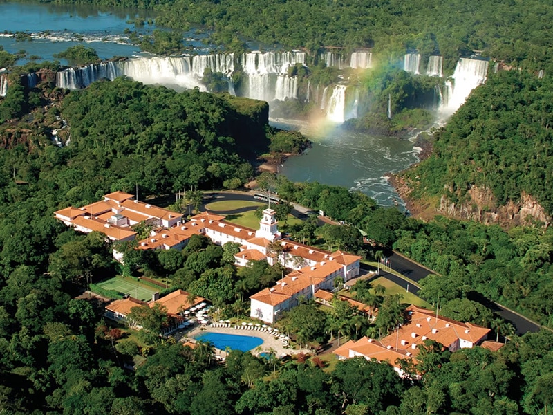 Vista aérea de um hotel em Foz do Iguaçu, cercado por vegetação e com as Cataratas do Iguaçu ao fundo, formando um arco-íris. O hotel possui telhados vermelhos e uma piscina.
