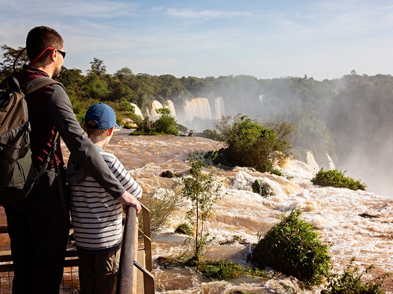 Pai e filho observando o cenário exuberante das Cataratas do Iguaçu, com a água caindo e a vegetação ao redor, enquanto desfrutam de um passeio na natureza.