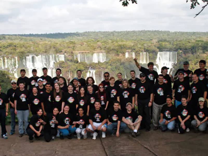 Grupo de pessoas vestindo camisetas pretas com o logotipo de "Loumar Turismo" posando em frente às Cataratas do Iguaçu, em um cenário natural exuberante.