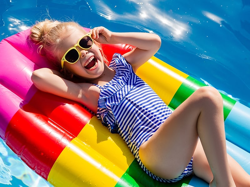 Menina sorridente de óculos de sol e maiô listrado, relaxando em uma boia colorida na piscina em um dia ensolarado.