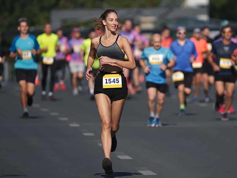 Mulher participando de uma corrida de rua em Foz do Iguaçu, usando um top cinza escuro e shorts pretos, com o número de inscrição 1545 preso à frente. Ela está correndo em uma avenida larga, acompanhada por muitos outros corredores, todos vestindo roupas esportivas coloridas. A expressão determinada da mulher destaca seu foco e energia durante o evento. As corridas de rua em foz do iguaçu acontecem em um dia ensolarado, proporcionando um cenário vibrante e dinâmico para a competição.