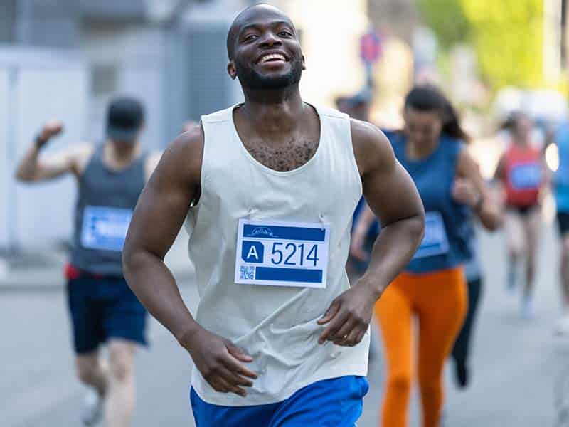 Homem sorridente correndo em uma corrida de rua, vestindo uma regata branca e shorts azuis, com o número de inscrição A 5214 preso à frente. Outros corredores são visíveis ao fundo, participando do mesmo evento. A corrida acontece em um ambiente urbano, com prédios e árvores ao longo do percurso. A expressão de felicidade do corredor em destaque sugere um momento de superação e alegria durante a prova.