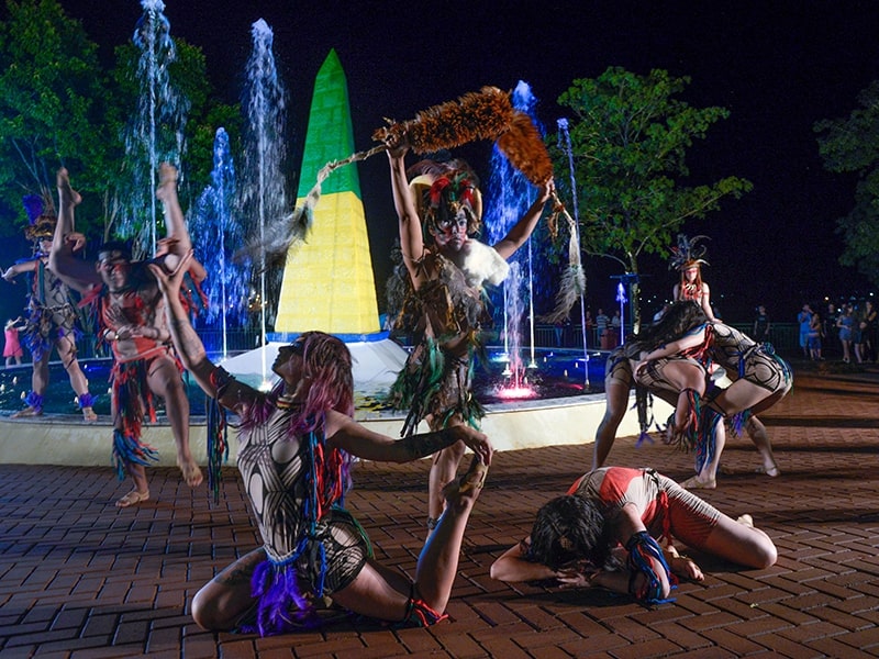 Apresentação noturna de dança tradicional em frente a uma fonte iluminada em Foz do Iguaçu. Os dançarinos estão vestidos com trajes indígenas coloridos e adornos, realizando movimentos expressivos e coreografias dinâmicas. Ao fundo, a fonte iluminada com cores vibrantes e uma grande escultura em forma de cone adicionam um elemento visual impressionante à cena. Este evento cultural destaca a rica herança e tradições da região, proporcionando uma experiência única e envolvente para os espectadores.