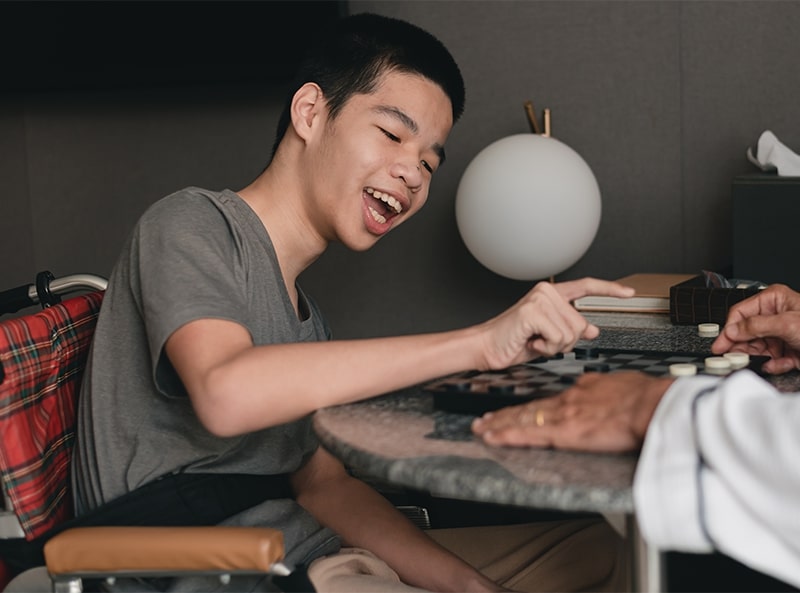 Quartos para Portadores de Necessidades Especiais - Imagem de um jovem em cadeira de rodas sorrindo enquanto joga um jogo de tabuleiro com outra pessoa em um quarto de hotel, destacando a acessibilidade e o ambiente acolhedor.