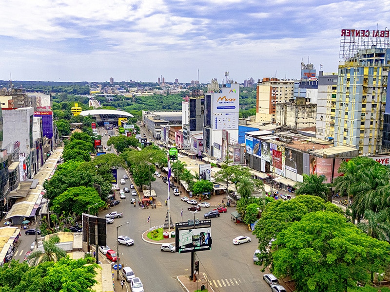 Vista aérea do centro de Ciudad del Este, no Paraguai, com diversas lojas e prédios comerciais ao longo da avenida principal, rodeada por vegetação exuberante. A imagem destaca a movimentação típica da cidade, que é um dos principais destinos para quem procura onde comprar Lego no Paraguai, com várias opções de lojas especializadas em brinquedos e eletrônicos.