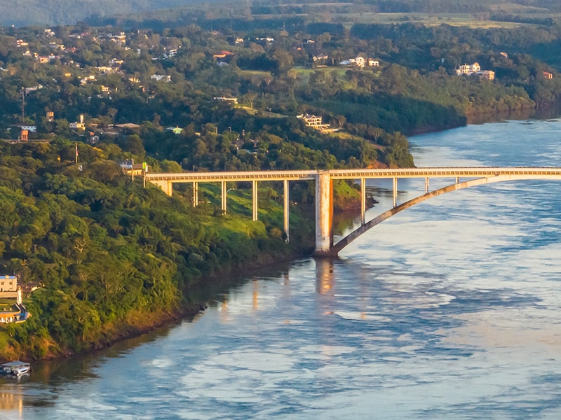 Vista aérea da Ponte Internacional da Amizade sobre o Rio Paraná, ligando Foz do Iguaçu, Brasil, a Ciudad del Este, Paraguai. A ponte é cercada por vegetação densa e construções espalhadas, com o rio refletindo a luz do sol.