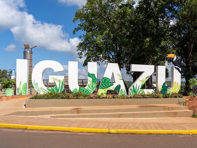 Placa decorativa com a palavra 'IGUAZU', decorada com imagens de folhas tropicais e um tucano, localizada em uma área ao ar livre, com árvores ao fundo e céu azul.