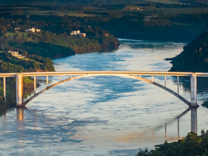 Ponte rodoviária cruzando um amplo rio cercado por vegetação densa em ambos os lados. O reflexo da estrutura é visível na água calma ao entardecer. Cenário típico de fronteira na América do Sul.