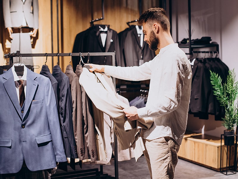 Homem em uma loja de roupas masculinas, examinando um terno bege enquanto está cercado por diversos ternos e jaquetas pendurados. O ambiente da loja é elegante e bem iluminado, com uma planta decorativa ao fundo.