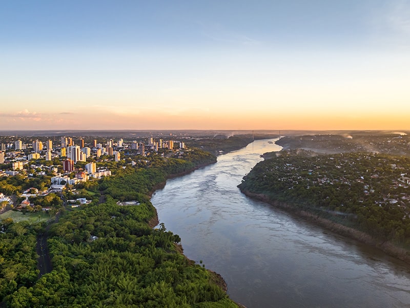 Vista aérea do Rio Paraná ao pôr do sol, separando as cidades de Foz do Iguaçu e Ciudad del Este, próximo ao Shopping China, rodeado por áreas urbanas e vegetação exuberante.