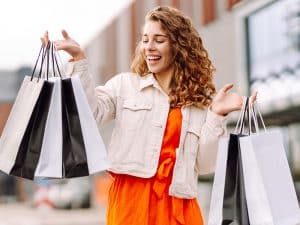 Mulher feliz fazendo compras ao ar livre, segurando várias sacolas de lojas de roupas. Ela está vestida com um vestido laranja vibrante e jaqueta clara, sorrindo enquanto caminha. Imagem relacionada às melhores lojas de roupas no Paraguai.