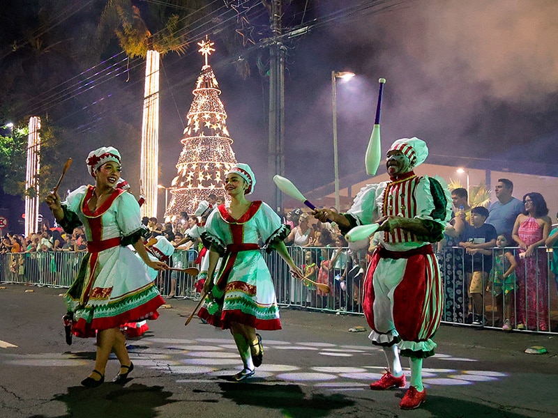 Artistas vestidos com trajes tradicionais dançam em um desfile natalino ao ar livre, com uma árvore de Natal iluminada ao fundo e espectadores observando ao longo da rua.