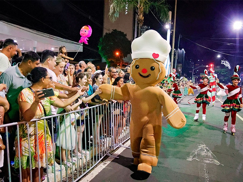 Personagem de biscoito de gengibre interage com o público durante desfile de Natal em Foz do Iguaçu, em uma rua iluminada e cheia de espectadores, criando uma atmosfera festiva e alegre.