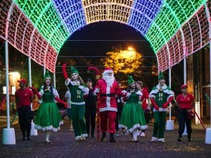 Desfile de Natal em Foz do Iguaçu com Papai Noel e ajudantes vestidos de verde sob um túnel iluminado com luzes coloridas, criando um ambiente festivo e encantador.