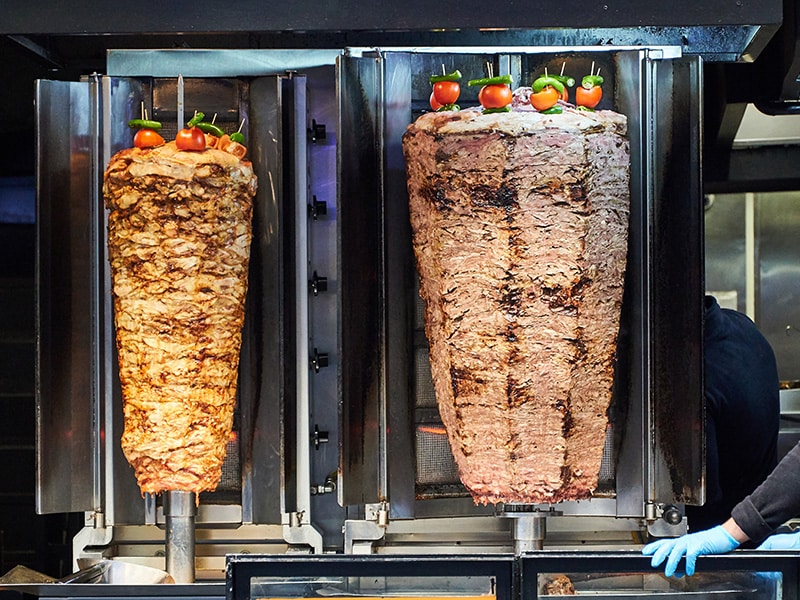 Dois espetos de shawarma com carne grelhando, decorados com tomates, prontos para serem servidos em um restaurante.