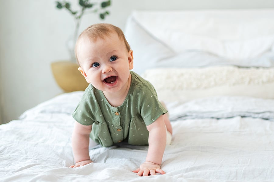 Bebê sorrindo deitado em uma cama, vestindo um macacão verde.