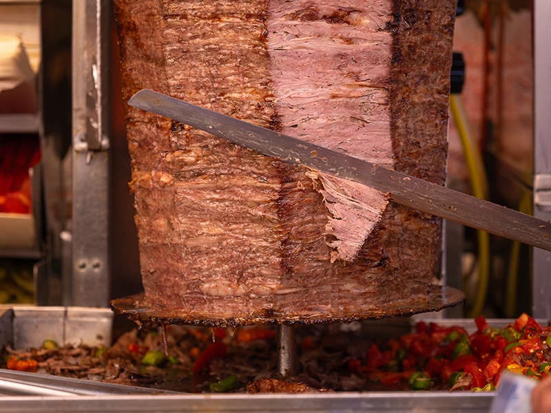 Carne de shawarma sendo cortada em um espeto giratório, pronta para ser servida em um prato tradicional.