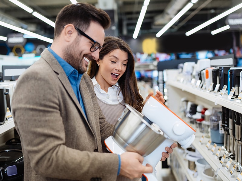 Casal animado explorando eletrodomésticos em uma seção de utensílios de cozinha no Shopping China, examinando uma batedeira com entusiasmo. Imagem ilustrativa.