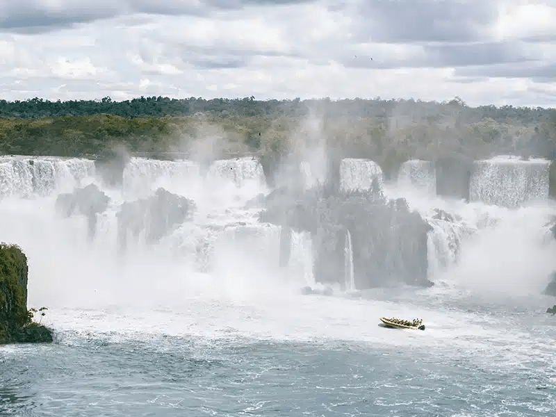 Vista panorâmica das Cataratas do Iguaçu, com várias quedas d'água caindo em meio a uma densa neblina criada pelo impacto da água. Um barco com turistas navega nas águas turbulentas perto das quedas, destacando-se em meio à grandeza das cataratas. Ao fundo, uma vegetação densa cobre as margens, enquanto nuvens cinzas se espalham pelo céu, criando um cenário dramático e impressionante.