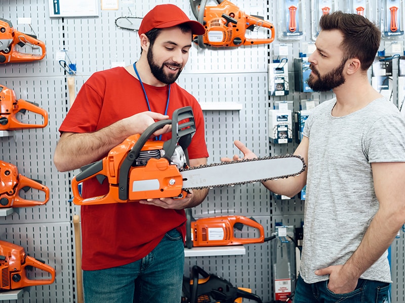 Vendedor explicando as funcionalidades de uma motosserra elétrica para um cliente em uma loja especializada em ferramentas elétricas. A imagem mostra uma variedade de equipamentos expostos, com destaque para a qualidade e a diversidade de produtos disponíveis para profissionais e amadores.