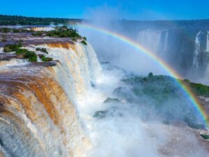 Vista das Cataratas do Iguaçu, um dos melhores lugares para viajar, com quedas d'água majestosas e um arco-íris brilhante formando-se sobre o rio, em meio a uma paisagem verde exuberante e céu claro.