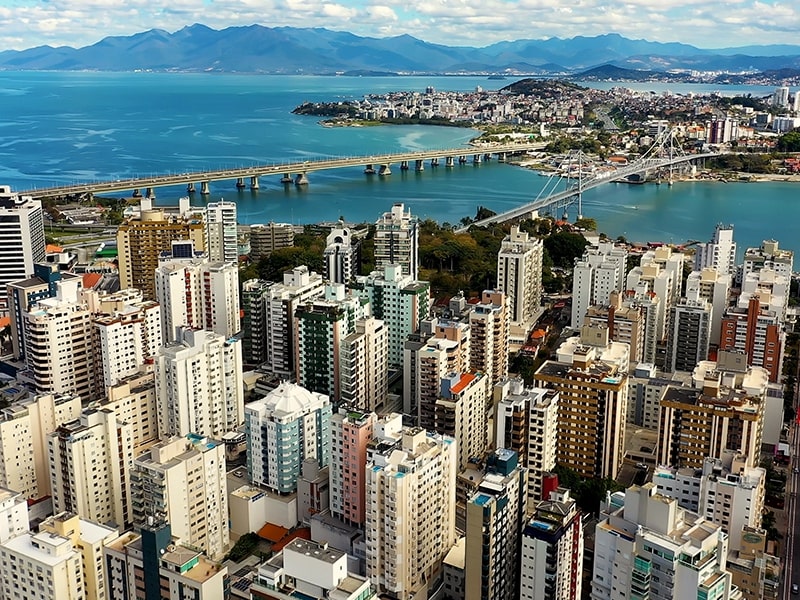 Vista aérea de uma cidade litorânea com muitos edifícios altos, uma ponte conectando diferentes áreas sobre o mar azul e montanhas ao fundo, formando uma paisagem urbana cercada pela natureza.