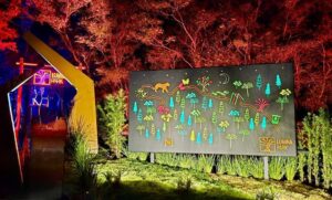 Painel iluminado e colorido no Lumina Park, exibindo uma cena vibrante de flora e fauna com árvores e animais estilizados sob um céu noturno. Ao fundo, um portal de luz brilhante em tons vermelhos e amarelos cria um ambiente mágico, destacando o cenário ao ar livre e trazendo uma atmosfera de encantamento ao local.