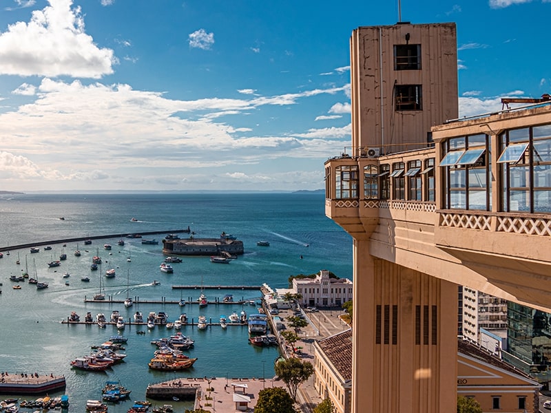 Vista do Elevador Lacerda em Salvador, Bahia, com o mar ao fundo, barcos ancorados na Baía de Todos-os-Santos e o Forte de São Marcelo em destaque, compondo um cenário histórico e marítimo em um dia ensolarado.